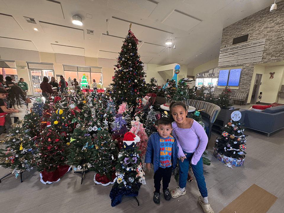 Two children smile in front of the mini Christmas trees in the great room at Ronald McDonald House San Diego